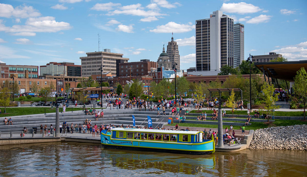 a passenger floater boat parked at the side of a river in a city park with the ity high rise buildings behind the people in park