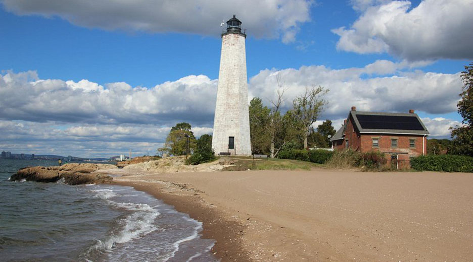 A white lighthouse in the center of a beach and red brick building to the right, calm ocean at left