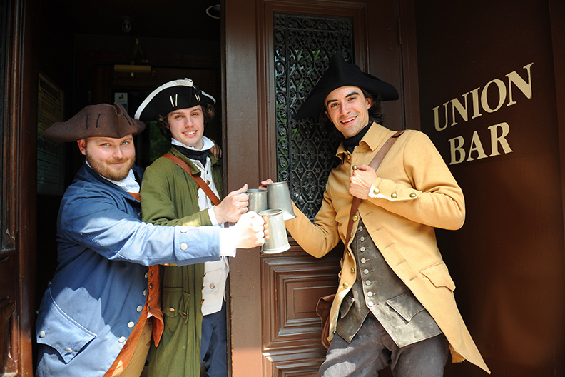 2 mwn toasting with beer mugs, dressed in 1700s era clothihng