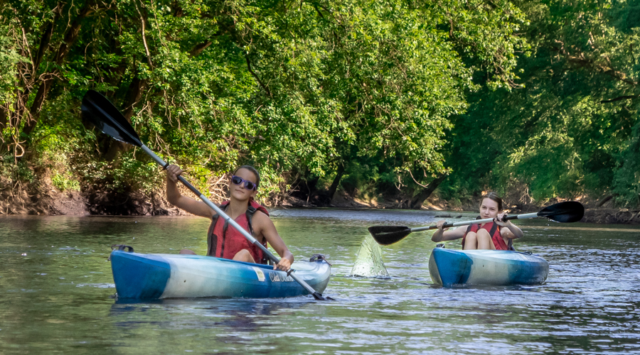 two kayakers in blue kayaks drifting down a river with trees in background 