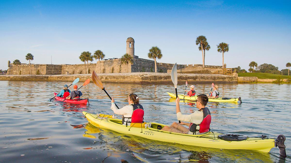 Kayaking people, in front of the old St. Augustine fortress. 2 kayaks are yellow; one is red. the fortress is made of bricks.