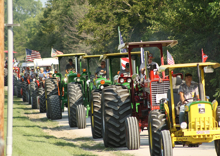 a parade of vintage tractors along the street, some green, the first one is yellow, second is red.