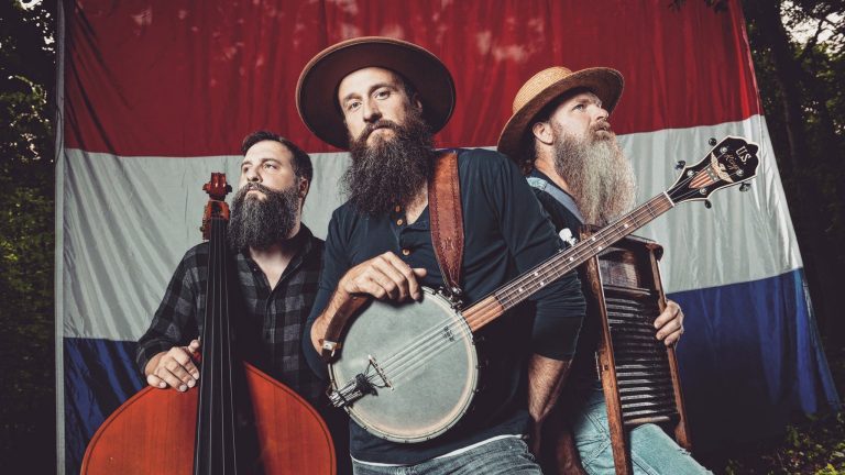 three men wearing beards, mustaches and wide-rim hats, standing with their musical instruments. The one standing in the center, is holding a bango, the one on right holds a washboard and the left one balances a bass fidle. 