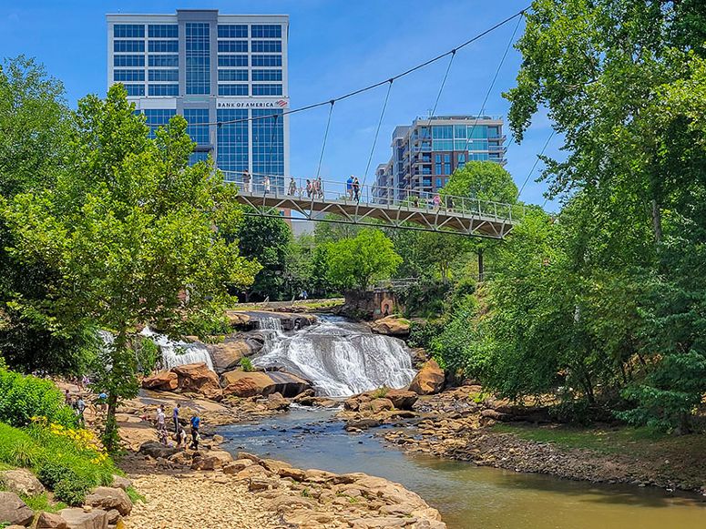 Falls Park on the Reedy. A pedestrian bridge over a small waterfall with green trees on both sides.