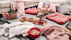 An indoor picnic. There is a tray of sandwiches along with a basket of wine and flowers on a blanket on the floor. They are surrounded by fluffy pillows and blankets.