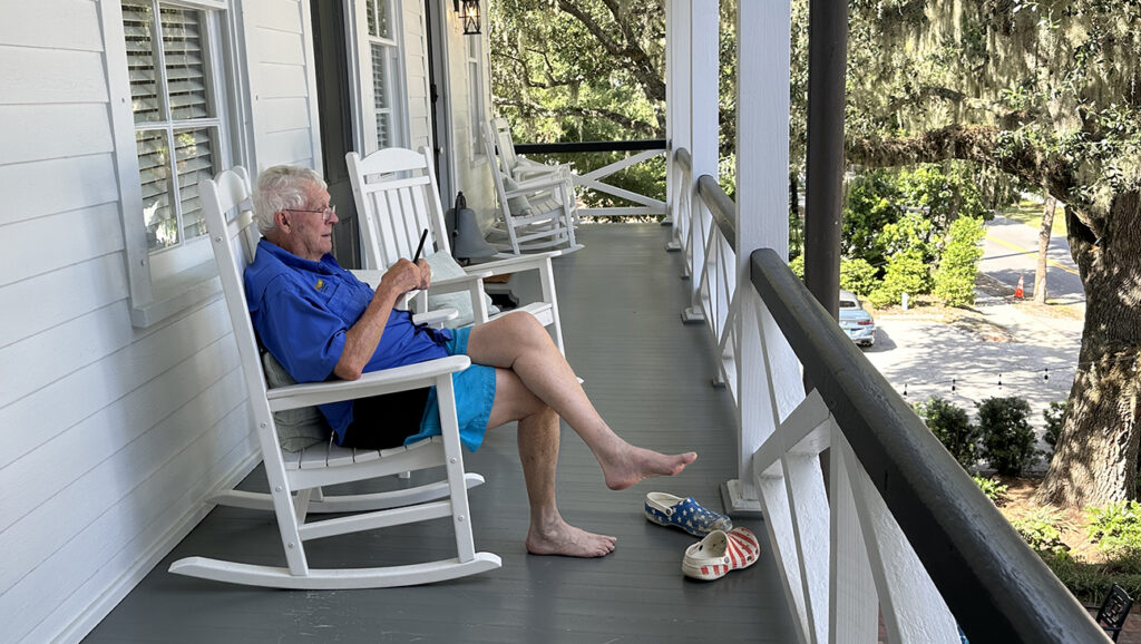 man sitting in a rocking chair on a wrap-around porch adn reading the paper