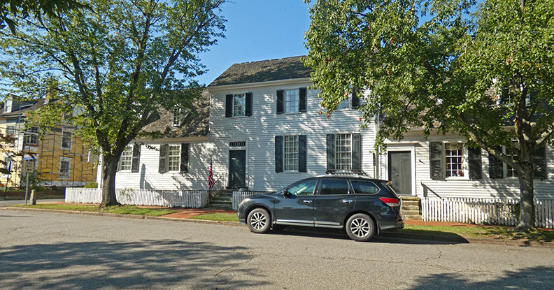 white 2-story colonial style house with black shutters on the two rows of windows. A black station wagon sits in front on the street. 