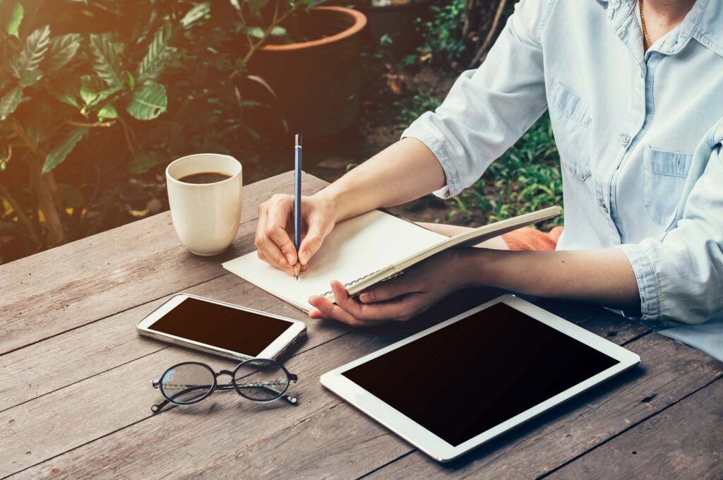writing notes in a notebook with a cell phone and tablet and cup of coffee on the table nearby