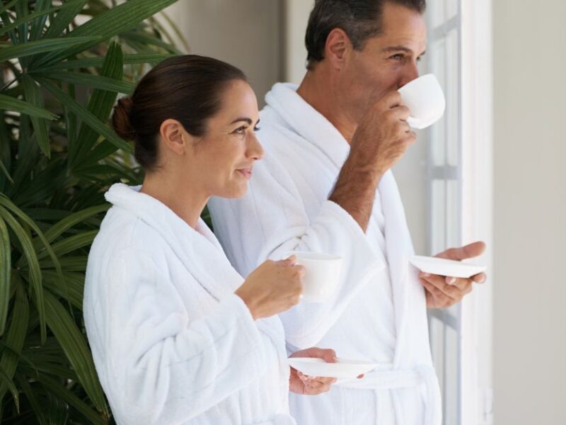 A couple in white robes looking out a window drinking coffee