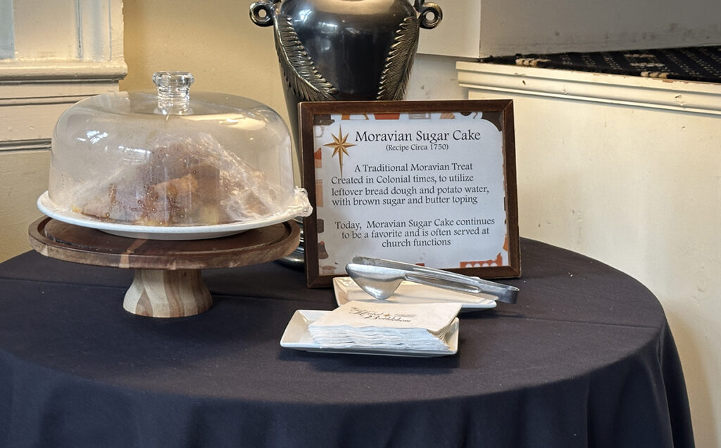 black round table set with a clear glass dome cake plate holding Moravian Sugar Cake and decorative sign describing it