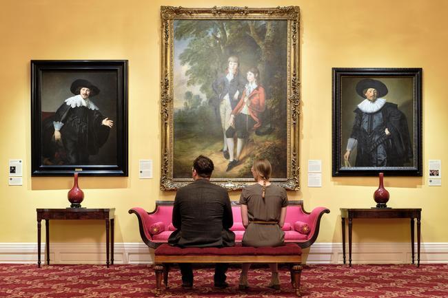 a couple siting on a pink couch with backs to camera and looking a 3 large paintings in frams on th wall. Th center is larger than the two on end. 