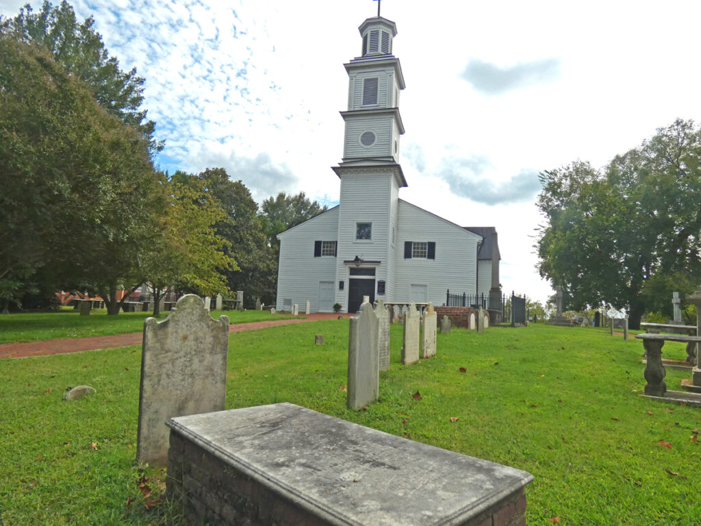 white church with a tall layered steeple and cemetery tombstones in front yard of church