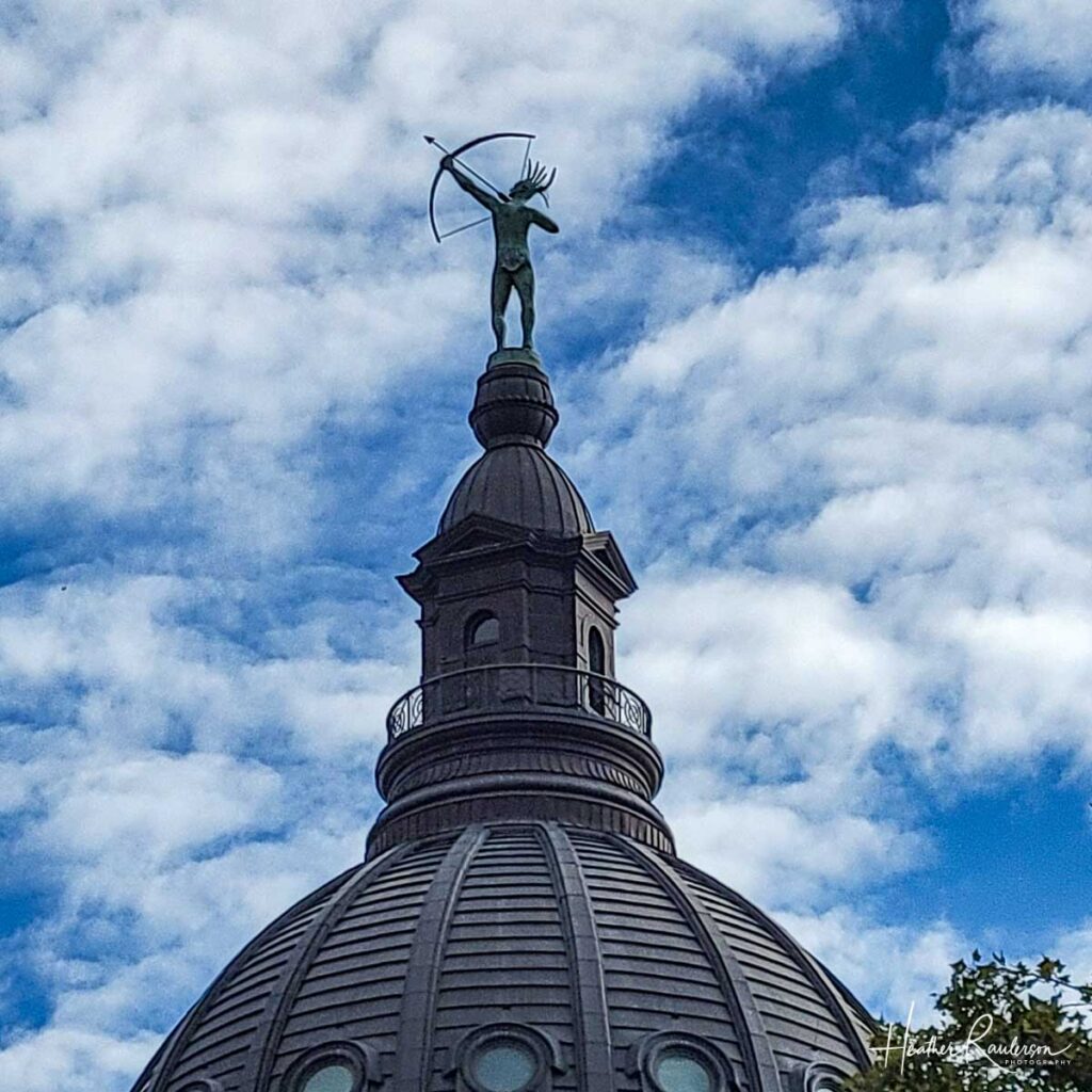 grey statue of indian holding bow and arrow on top of a round dome 