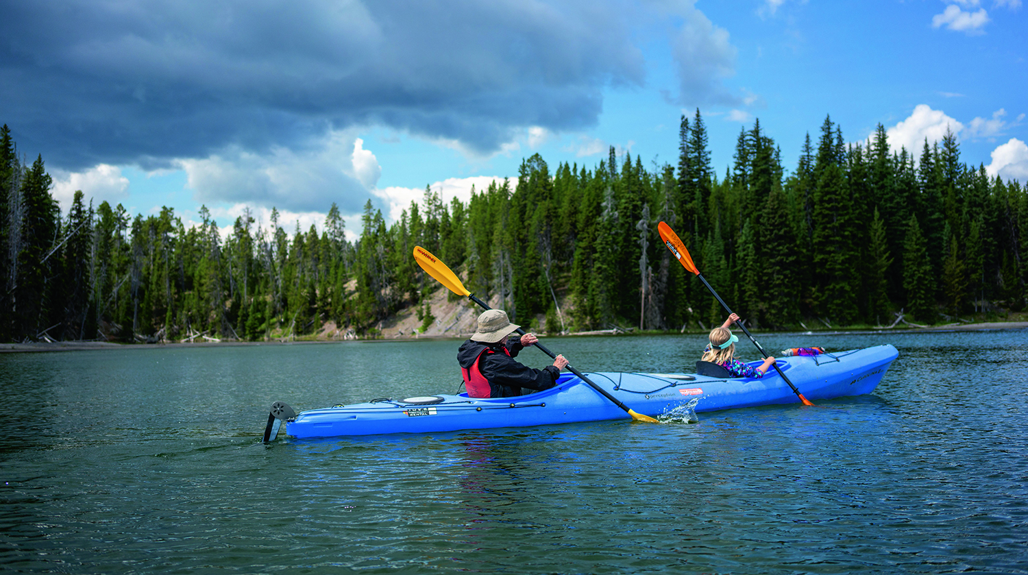 a couple canoeing and headed toward the right on a lake with trees behind them