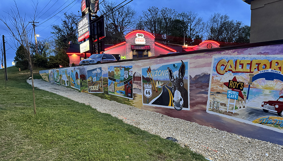 a long fence wall that has murals painted on it. Sidewalk runs along in front of it. Red roofs of buildings protrudebehind the wall
