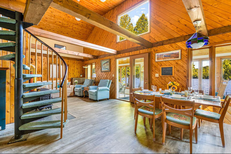 A pine wooden living room resort with high ceiling and picture window at top of peak, a curved staircase and dning table. 
