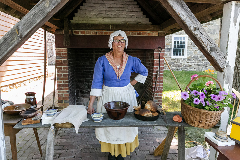 Lady dressed in colonial clothing standing behind a table of mixing bowls and food