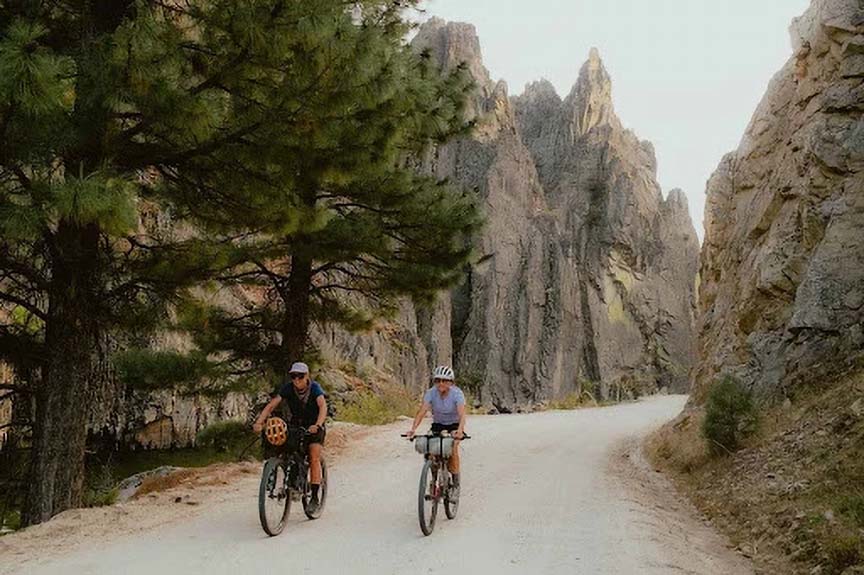 two bikers on a flat dirt road between hills of trees on both sides of road