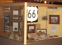 St.Rt. 66 sign on a post with memorabilia displayed on a table behind it. Wall is yellow