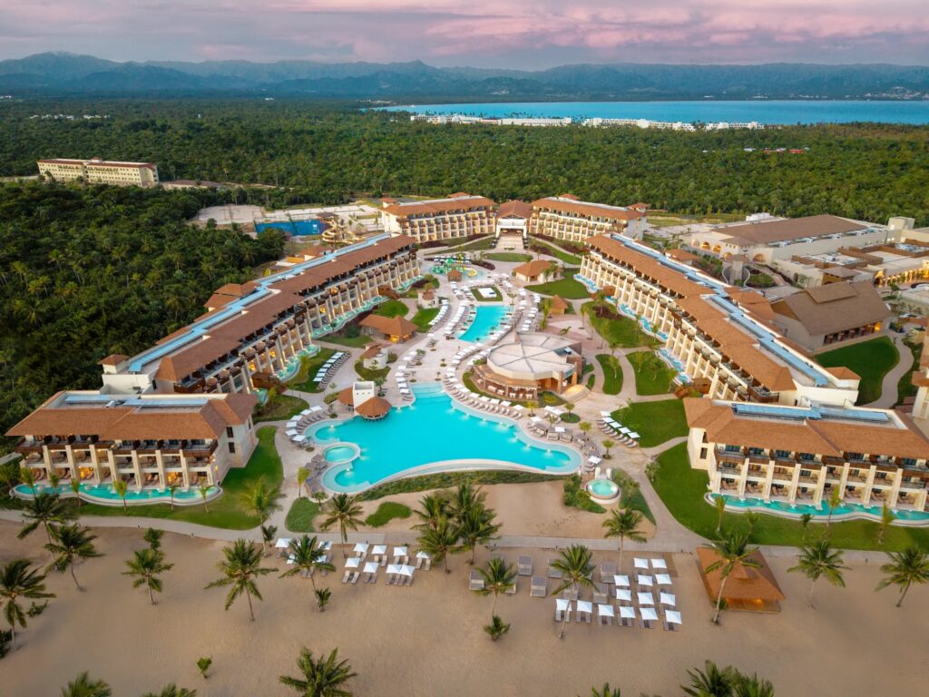 Areal view of a resort with 2-story buildings on both sides of pool in middle. The buildings then circle around the back of pool in an upside down U shape