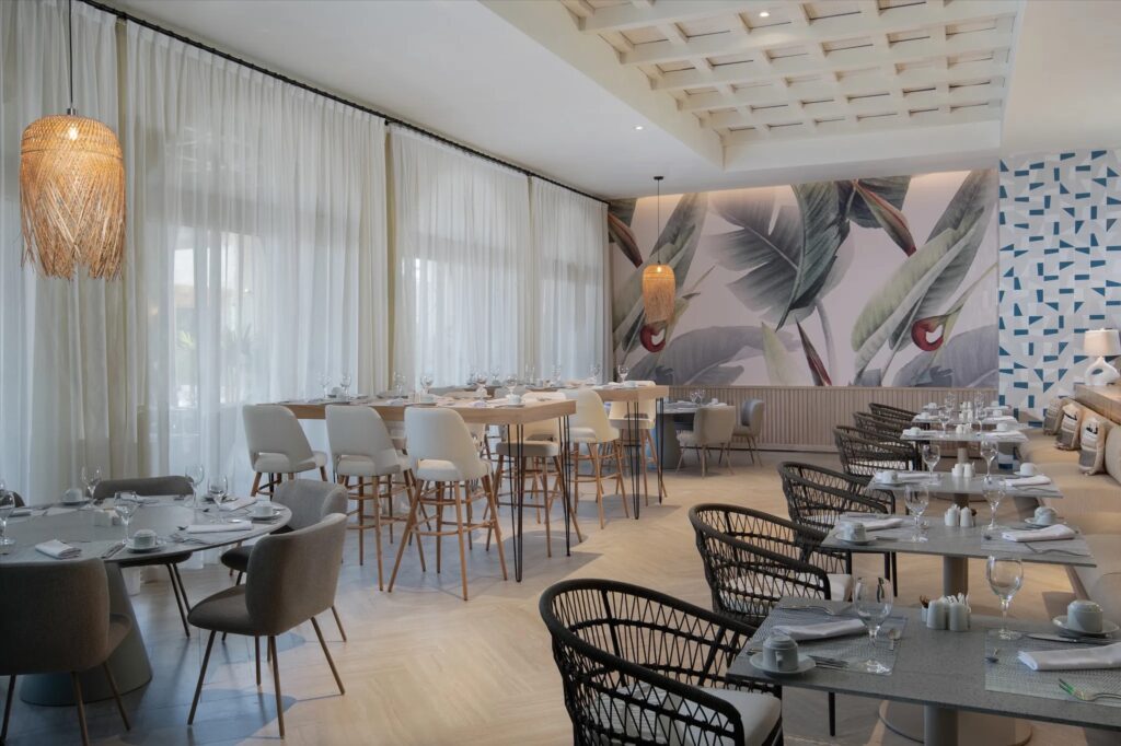 large modern dining cafe area with floor to ceiling windows and white sheer curtains. The back wall has a tropical modern tree leaves on it.