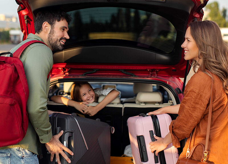 man and woman packing the car trunk with suitcases and daughter smiling and watching from the back seat