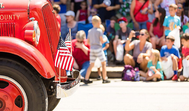 People along the street, sitting on the curb while watching a parade. At left, the front end of a red fire truck proceeds facing the right side.