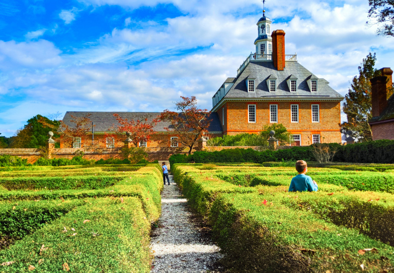 Historical colonial brick L-shapedbuilding with a grey roof with a chimney and a white window pillarat thetop. Trimmed bushes lie in frontof the building.