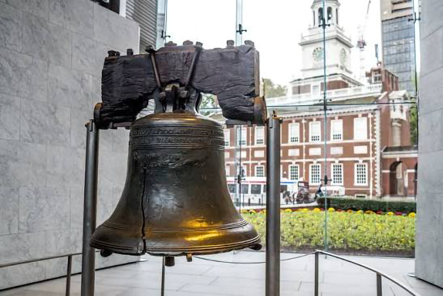 The U.S. Libertery bell mounted on its stand in front of a two-story colonial history building.