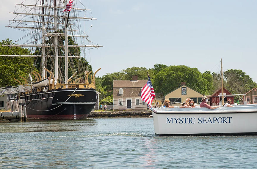 a huge sailing ship parked in water at left,  peaole sittingins an open air boat at rigt with a U.S. flag attached to the back end. Small budiness buildings inthe background at the water's  edge.