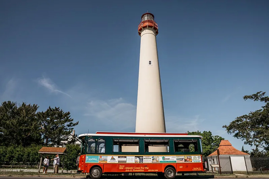 A red passenger tourist trolley parked in front of a tall white lighthouse. 