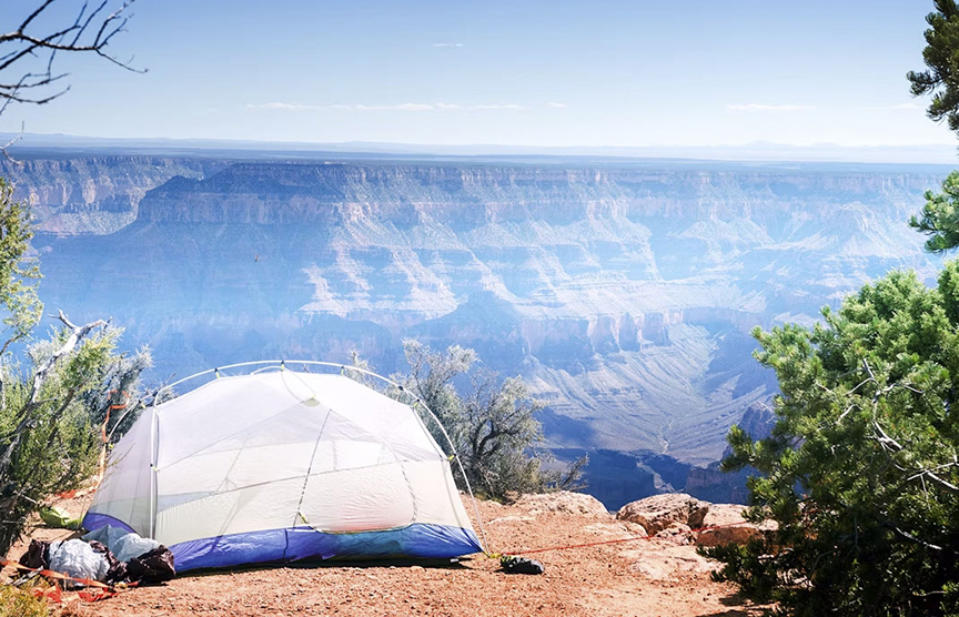 A white rounded tent set up at the edge of a cliff overlook blue hazy grand canyon mountains