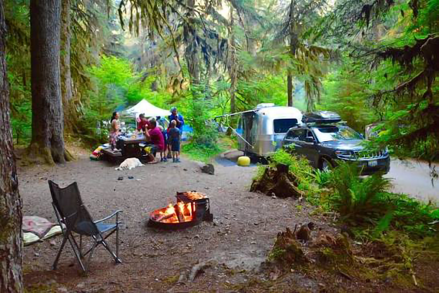 an rv camper to the right of a tent and acampfire in the middle of clearing. Trees in background
